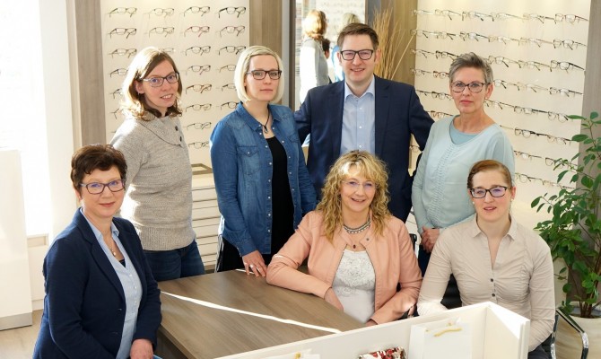 Team von Optikern steht in einem modernen Brillengeschäft vor einer Auswahl an Brillen.